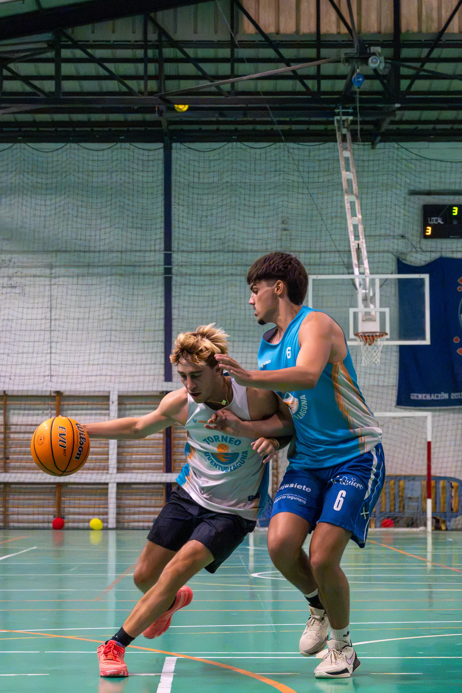 Jugador con camiseta blanca bota el balón ante un defensor con camiseta azul y el dorsal 6 en un pabellón