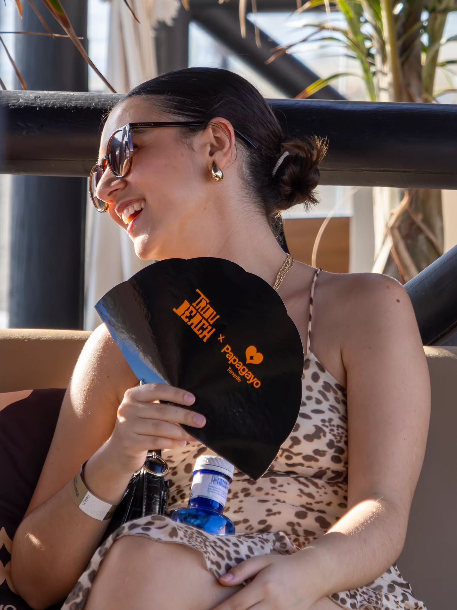 Chica sonriendo con gafas de sol y un abanico negro de Tribu Beach y Papagayo en un evento de día