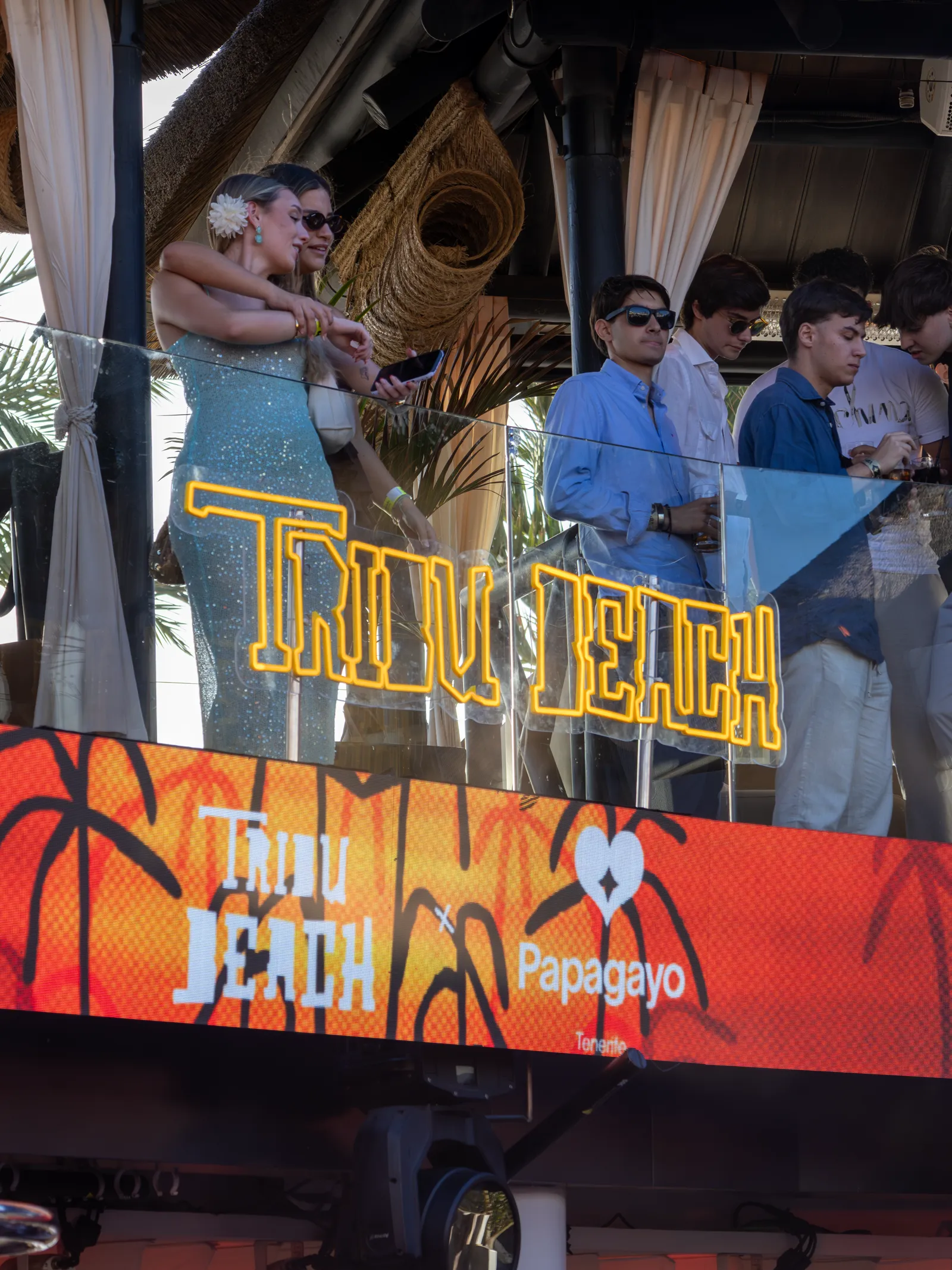 Público en el balcón de Papagayo Tenerife junto al neón amarillo de Tribu Beach y la pantalla del evento