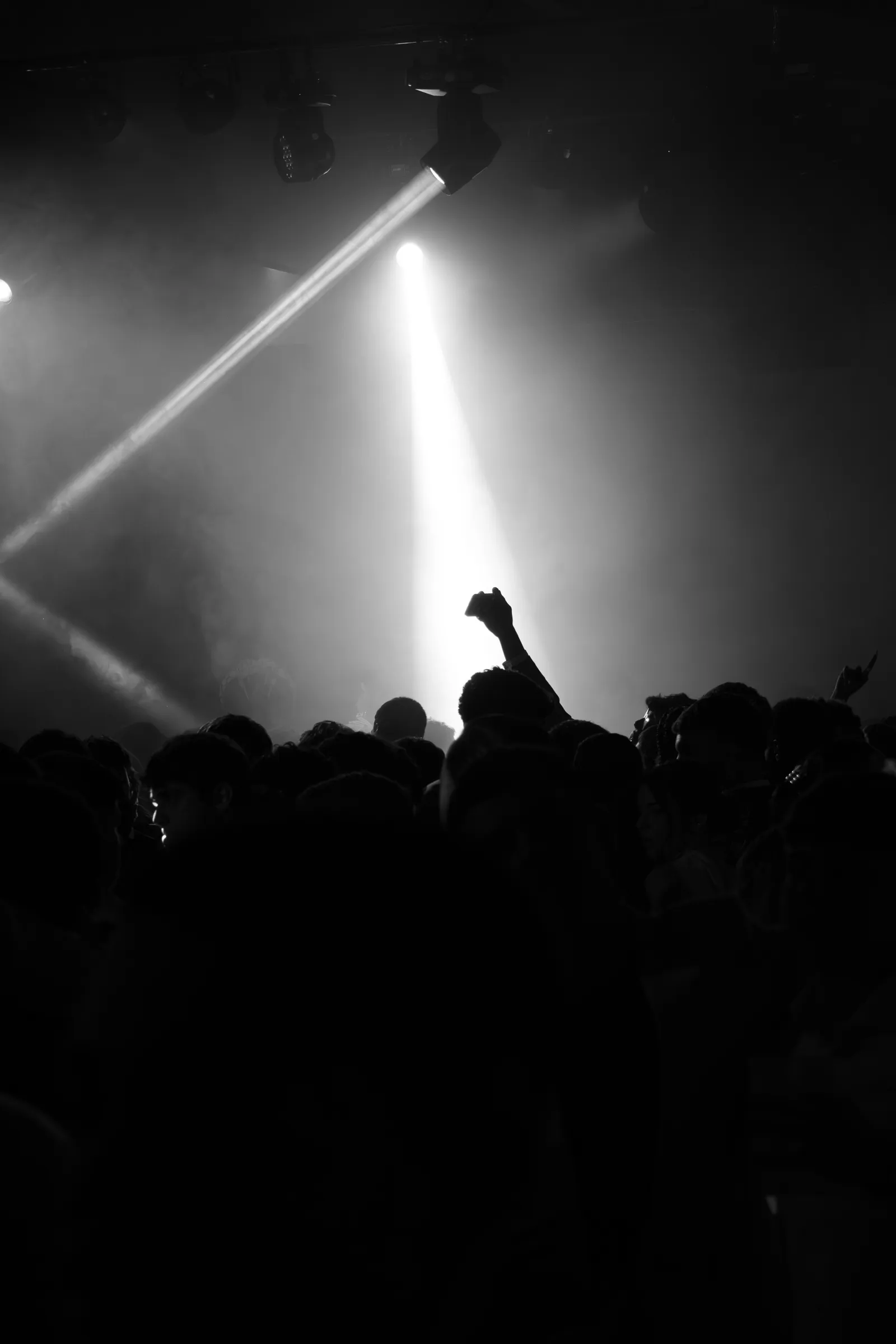 Siluetas del público con el brazo en alto bajo haces de luz entre el humo en The Last Nuit, en blanco y negro